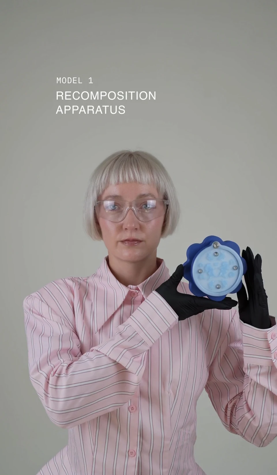 Nadia in a striped pink shirt and black gloves, presenting the blue Recomposition Apparatus.