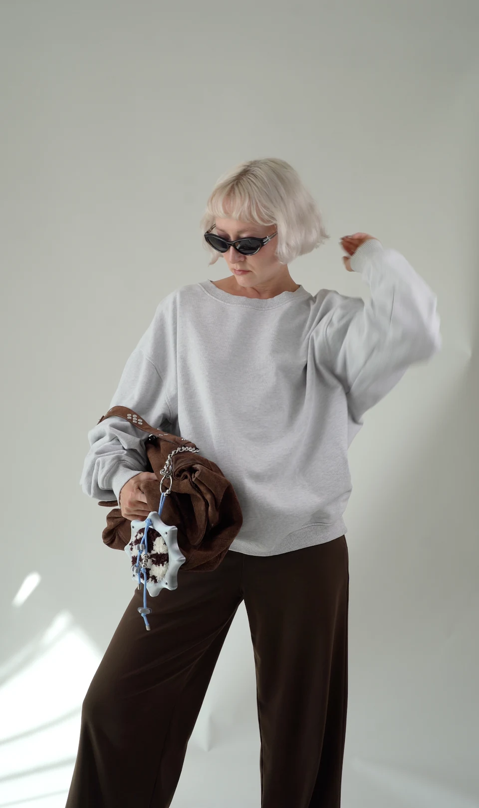 Nadia in a loose grey sweatshirt, brown trousers and sunglasses, wearing the tufted charm on her brown bag.
